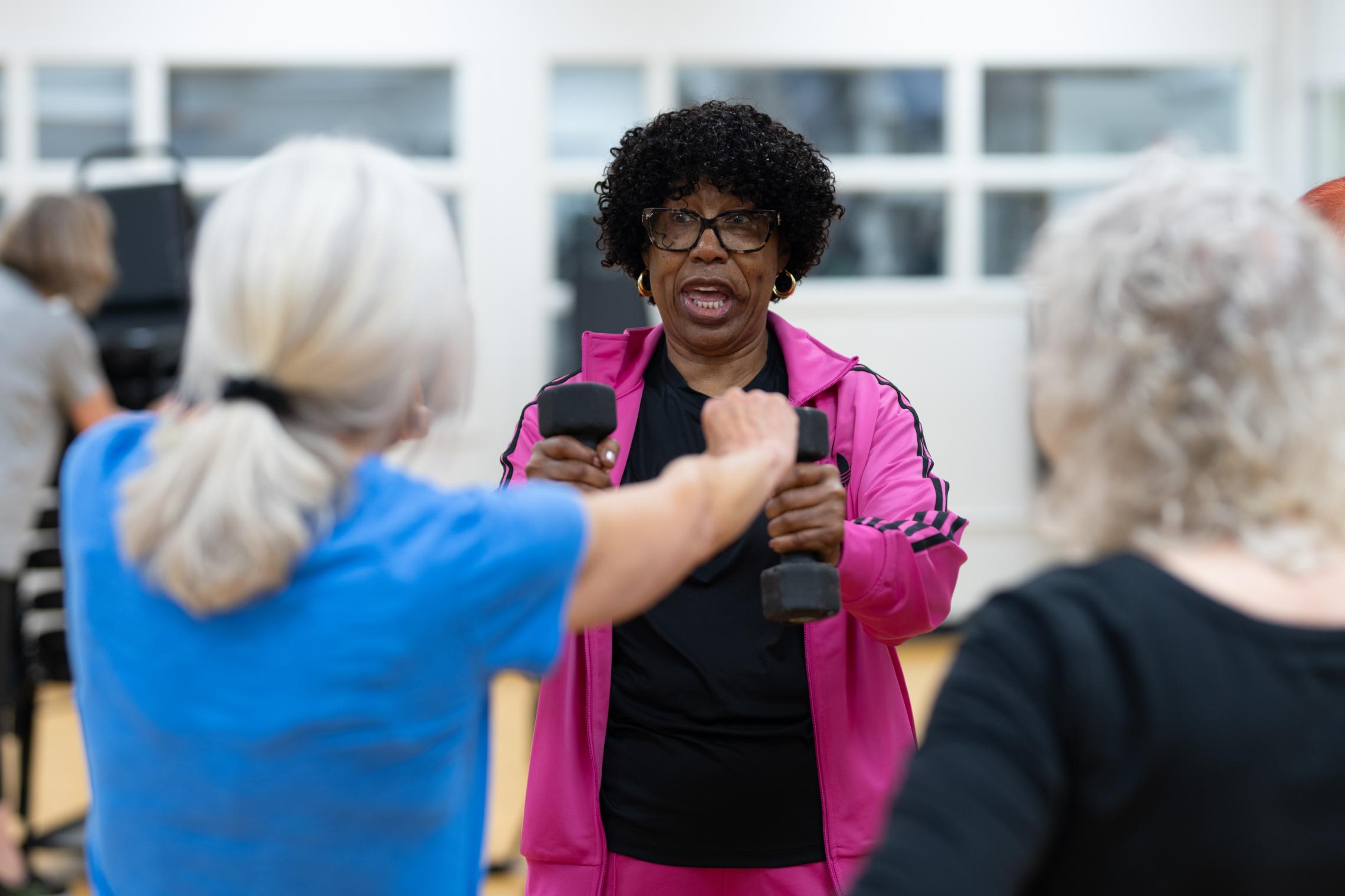 Participants in a YMCA group fitness class use hand weights while exercising in a gymnasium. One participant in a bright pink jacket lifts dumbbells in the center of the image as others follow along from chairs arranged throughout the room.