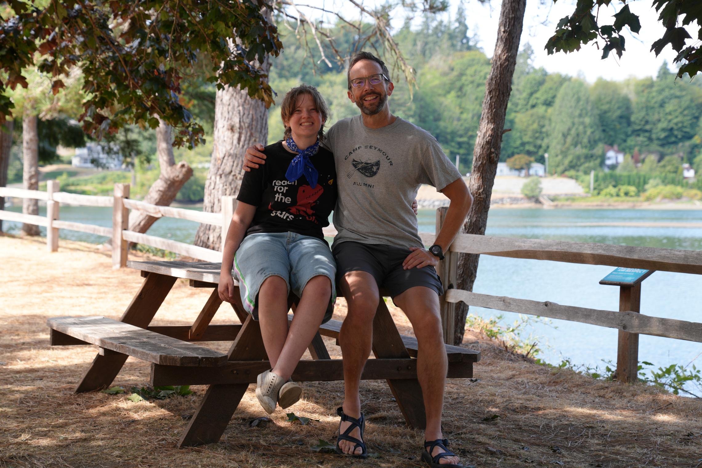 Alt text:  Aaron and Ryan sit together on a wooden picnic table at Camp Seymour beside the waterfront. Trees shade the area, with a wooden fence, calm water, and greenery visible in the background on a sunny summer day.