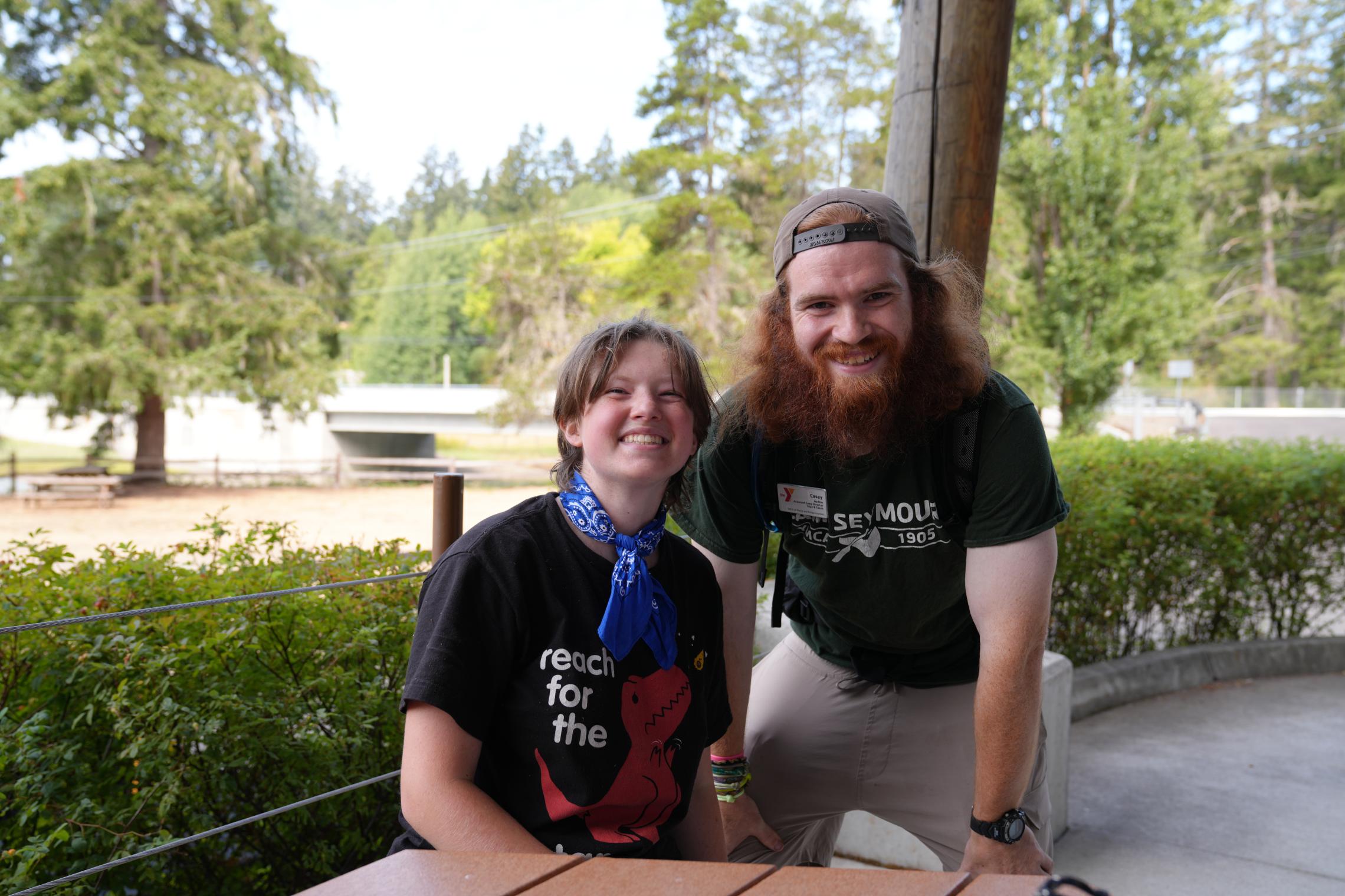 Ryan and Camp Seymour Assistant Camp Director Casey pose together at an outdoor gathering area. Ryan wears a blue bandana and camp T-shirt, while Casey wears a Camp Seymour shirt and name tag. Trees, shrubs, and camp grounds are visible in the background.
