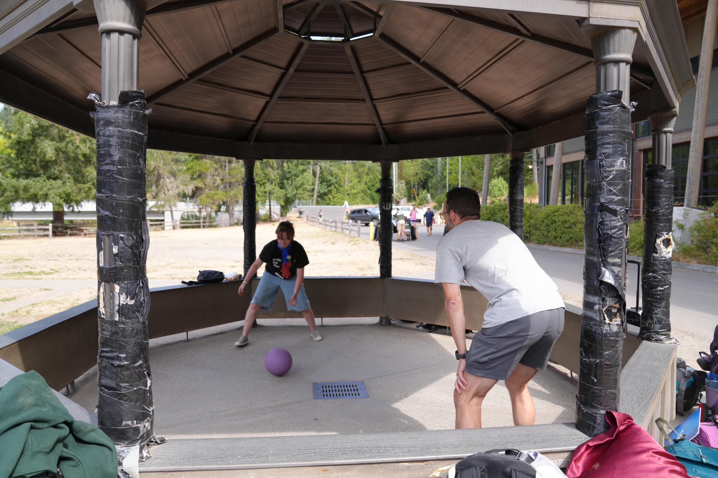 Aaron and Ryan play a game of gaga ball inside an octagonal gaga pit at Camp Seymour. A purple ball sits in the center as both players face each other, with camp buildings, trees, and open outdoor space visible in the background.