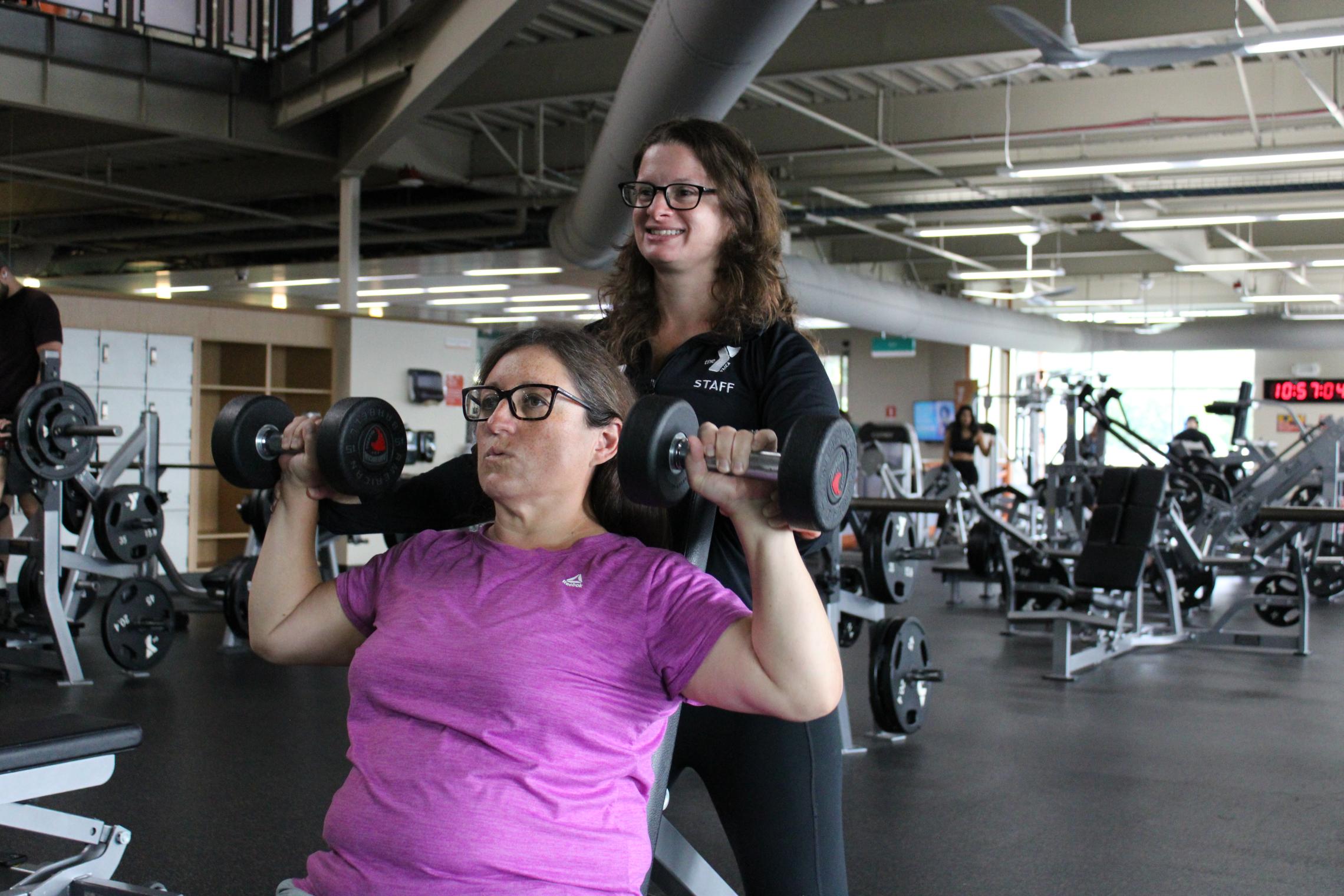 Julia performs a seated shoulder press with dumbbells at the Gordon Family YMCA while a YMCA staff member stands behind her for support. Weight machines, free weights, and other fitness equipment are visible throughout the wellness center.