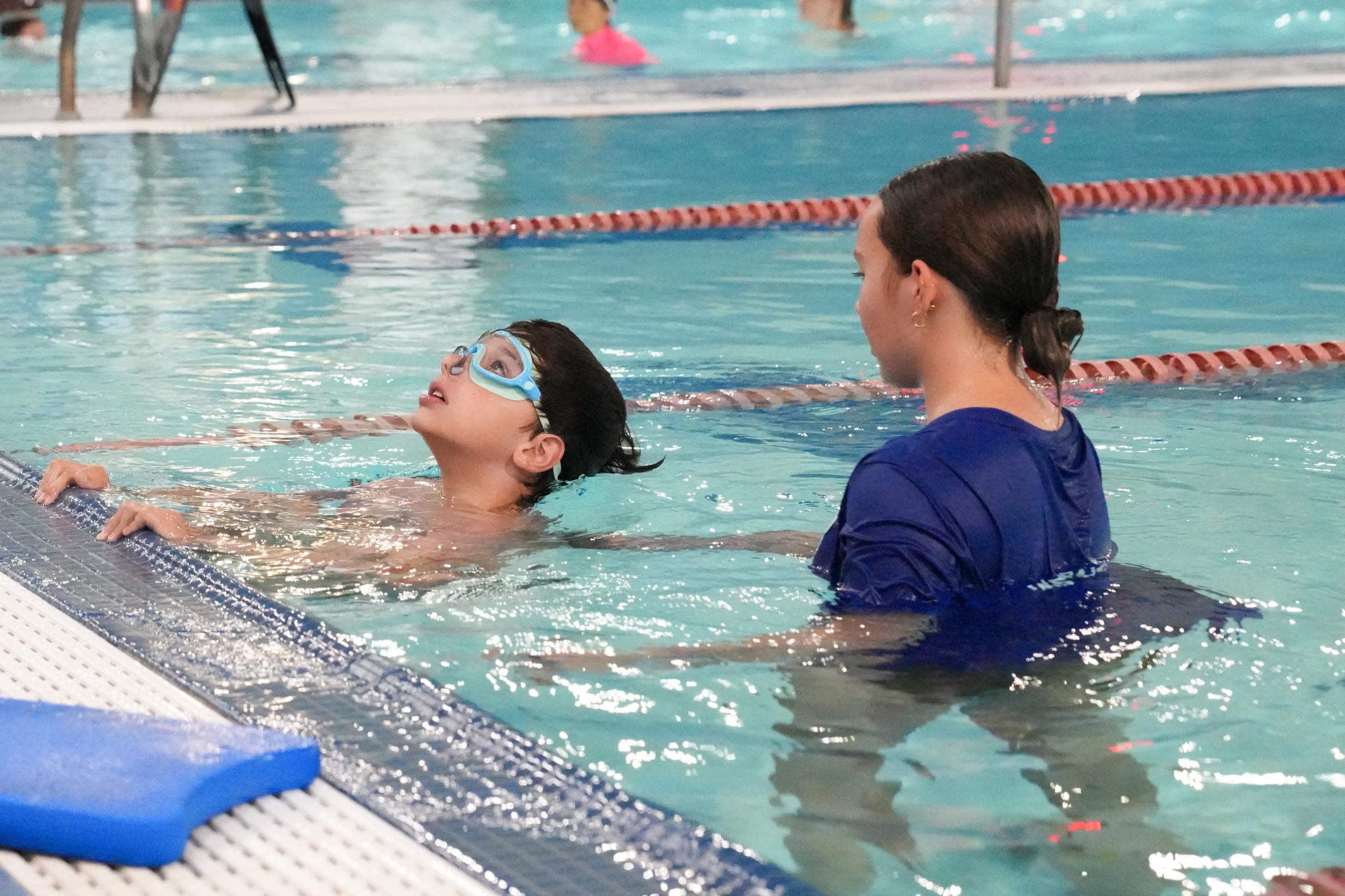 Child wearing swim goggles practices floating and swimming skills near the edge of an indoor YMCA pool during a private lesson. An instructor stands beside the child in the water, providing guidance, while lap lanes and other swimmers are visible in the background.