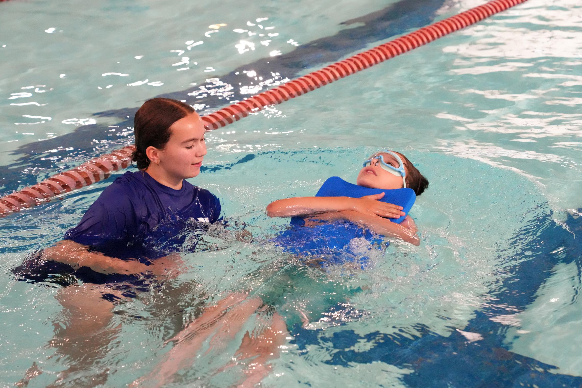 Child wearing swim goggles practices back floating with a blue flotation aid during a private swim lesson at an indoor YMCA pool. An instructor supports the child in the water nearby, while lane lines and clear pool water are visible in the background.
