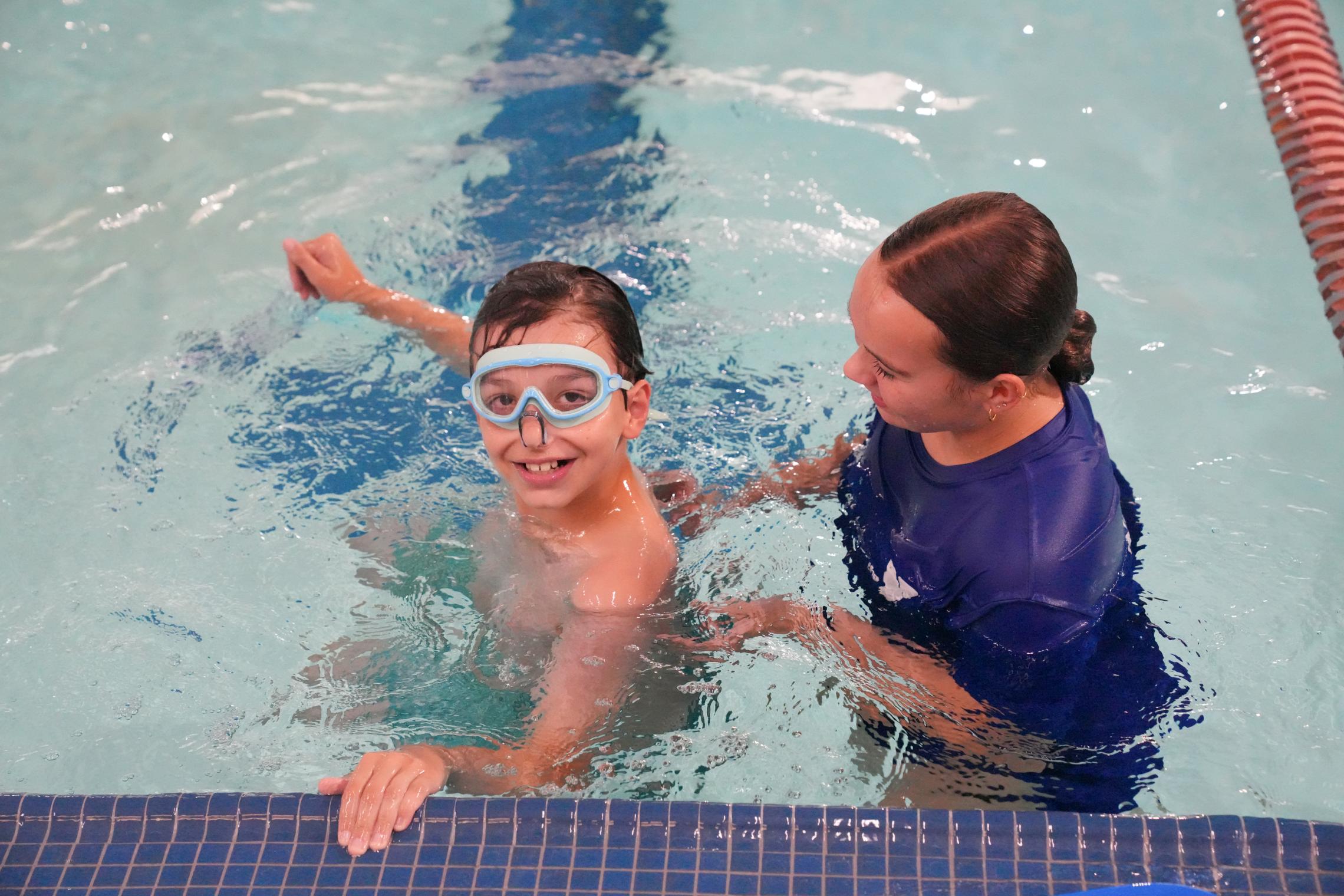 Child wearing swim goggles practices swimming independently near the edge of an indoor YMCA pool while an instructor provides support nearby in the water. Pool lane lines and clear blue water surround the lesson area.