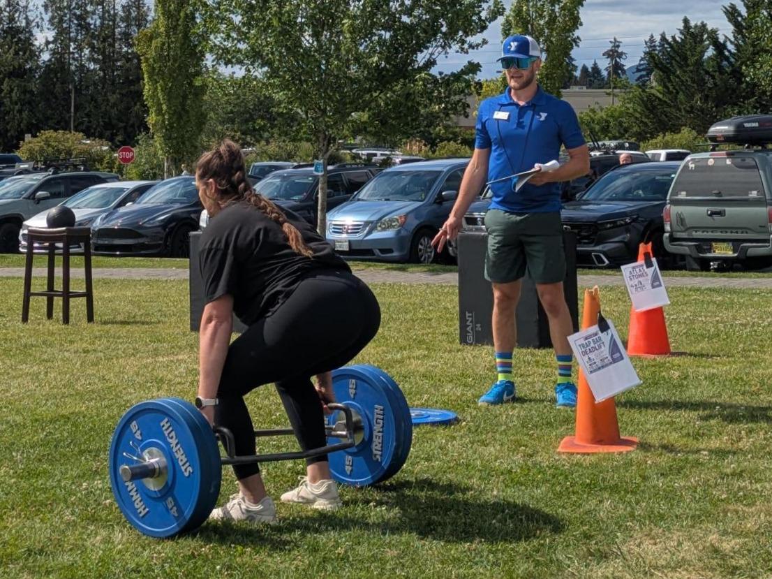 Participant performs a trap bar deadlift during the Haselwood Family YMCA Strongman Competition on the front lawn while a YMCA staff member monitors the event. Parked cars, event cones, and competition equipment are visible in the background.