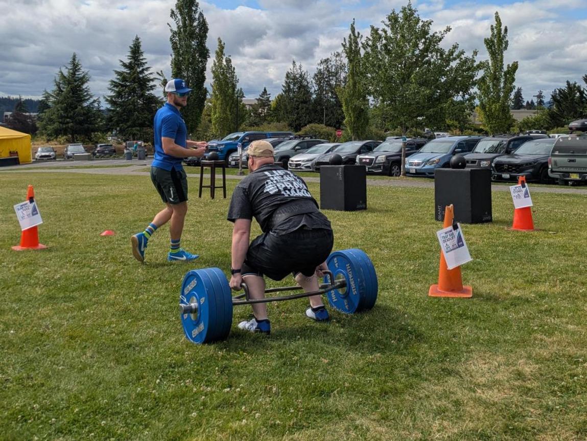 A participant performs a trap bar deadlift during the Haselwood Family YMCA Strongman Competition while an event official records results. Competition stations, cones, and strength equipment are set up across the YMCA front lawn.