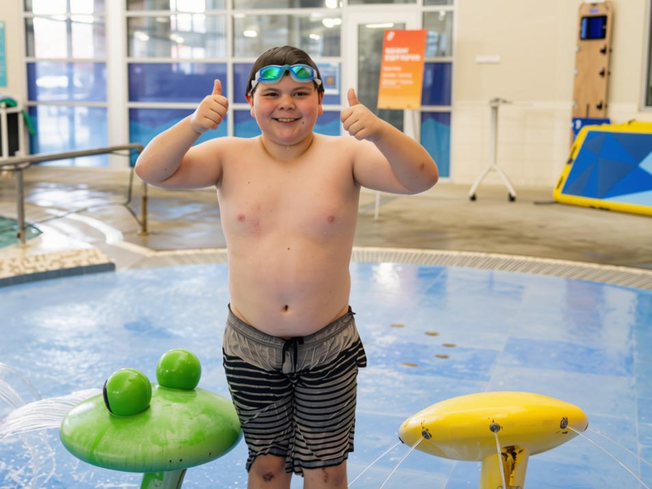 Beary gives two thumbs up while standing in the splash area at the Gordon Family YMCA after building confidence through YMCA swim lessons.