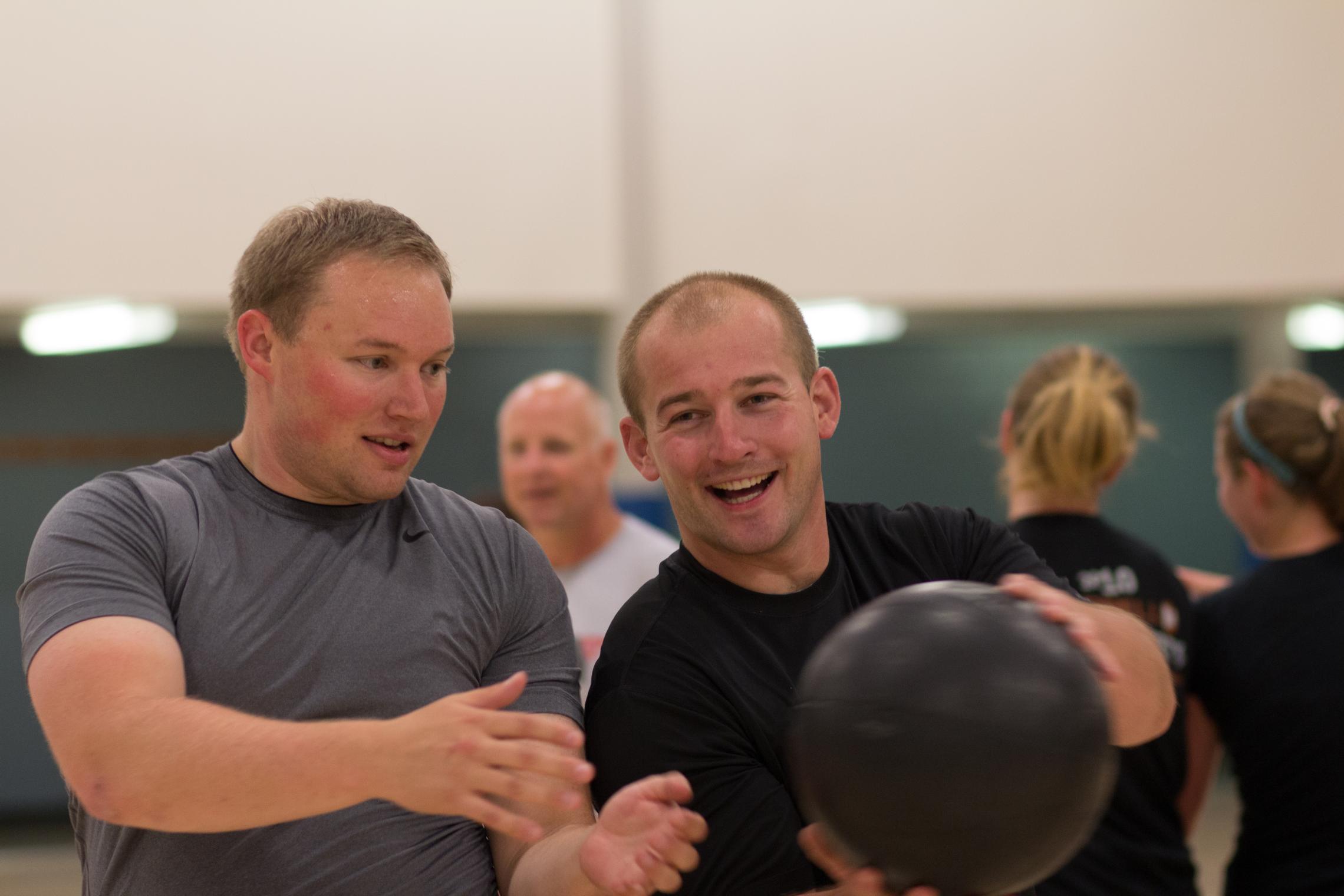 Two adults participate in a group fitness activity at a YMCA gym, passing a black medicine ball during a workout while other participants exercise in the background.