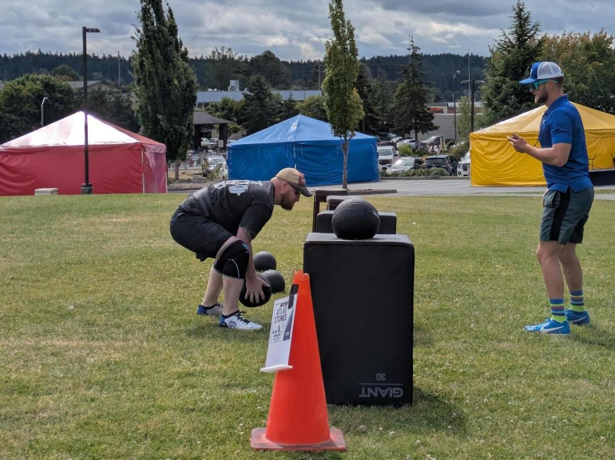 A competitor lifts an Atlas Stone during the Haselwood Family YMCA Strongman Competition while an event official tracks results. The outdoor strength challenge takes place on the YMCA front lawn with event equipment, cones, and colorful tents in the background.