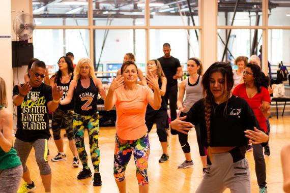 Dozens of people get into the groove of a Zumba class with their arms above and in front of them with various expressions of focus and glee. 
