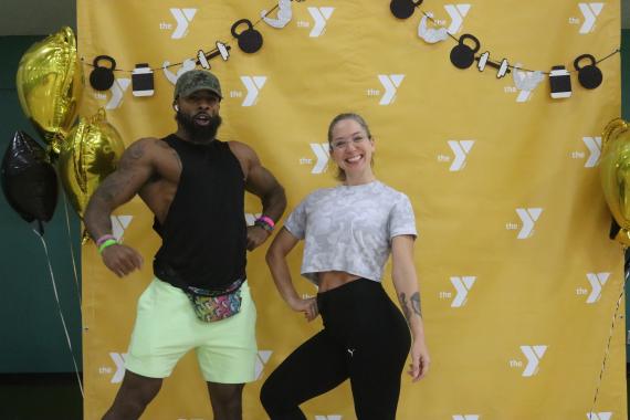 Fitness Fest attendees strike a strength pose in the photo booth area with a yellow step and repeat with the Y logo alongside gold and black balloons.