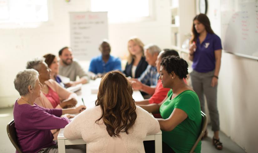 Diverse group of people sitting at a long table in the sunlight talking. Alongside them is a person near a white board with a YMCA polo shirt on.