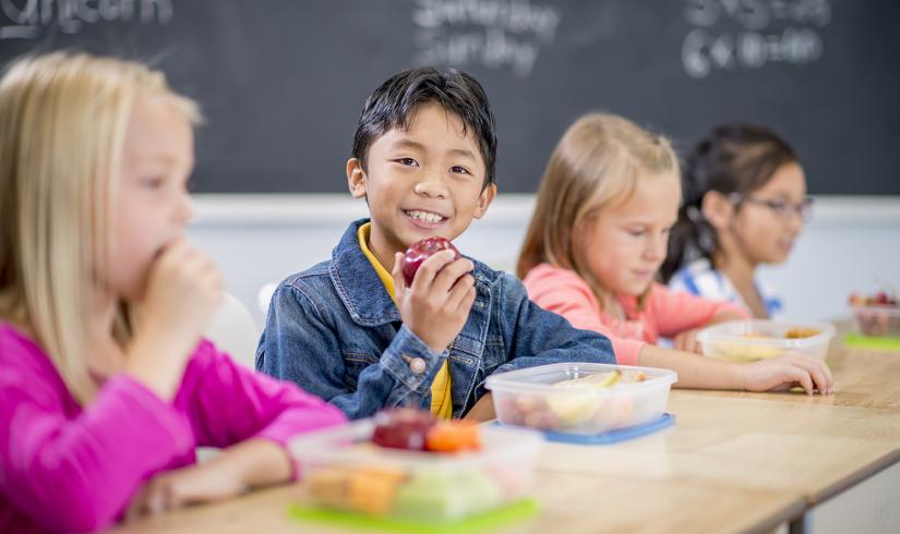 young boy smiling at the camera and holding an apple next to other young children sitting at a table