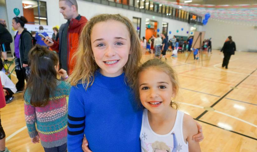 Two young girls stand side by side in a half hug within a glowing gymnasium. 