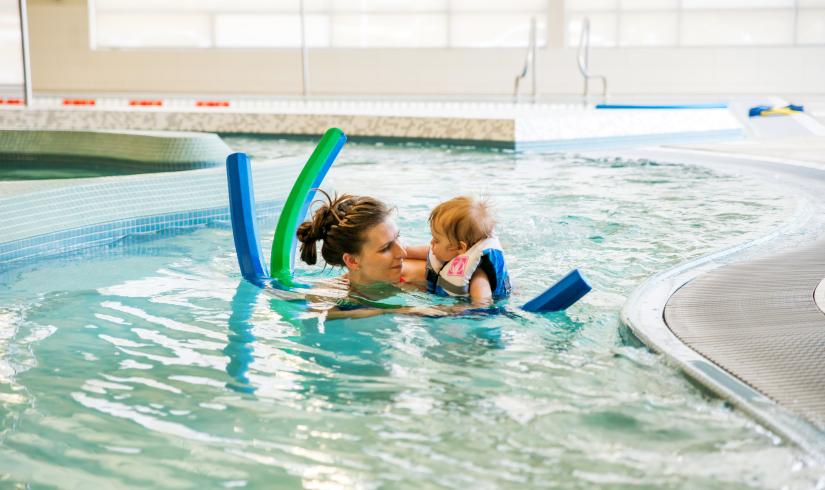 Woman holds a baby in the water. Baby is wearing a lifejacket and there are two pool noodles around them.