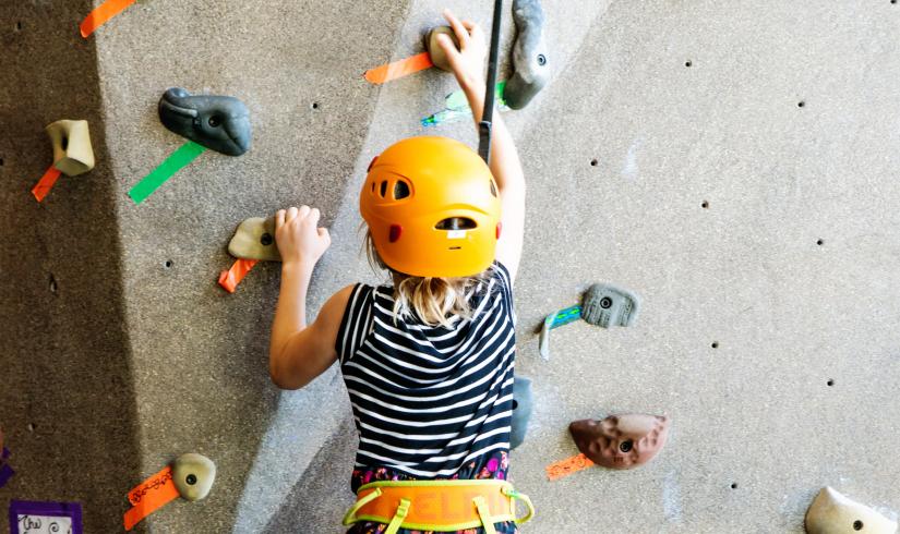 Kids is focusing climbing up a rock wall in an orange harness and helmet.