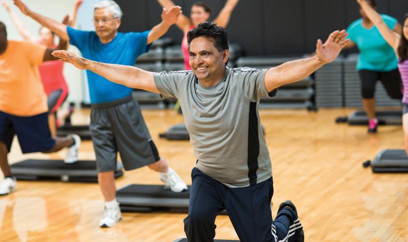 Man lunges on an aerobics floor with classmates. His arms extend into a Y shape and a smile is visible across his face.