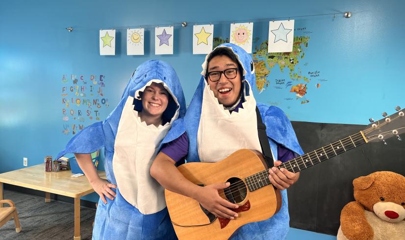 Instructors Lauren and Gabriel are in shark costumes and smiling as Gabriel holds an acoustic guitar.