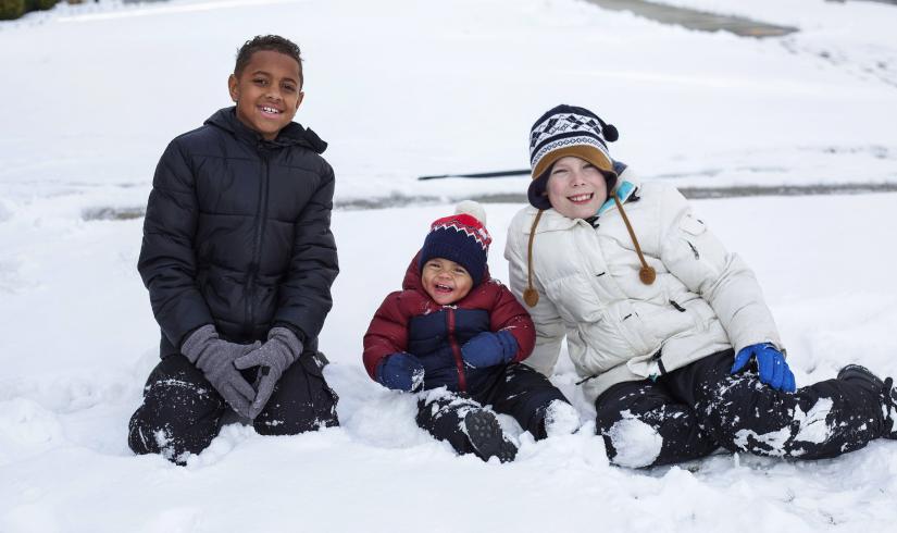 Three kids sit in the snow, two middle school age and one toddler.