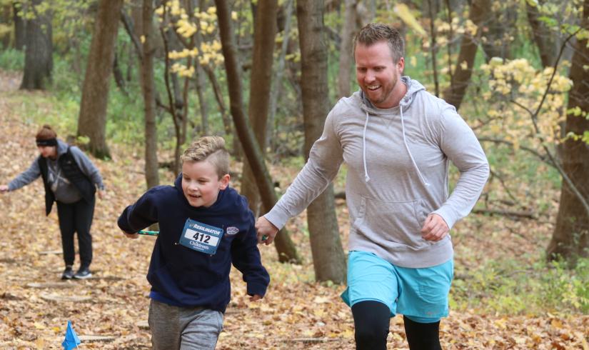 A father in son race up a trail, smiling as they move across fallen leaves on a cold day.