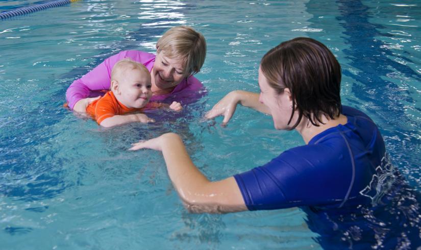 A guardian holds a baby in the water during an adult/child swim lesson as a Y staff member gently splashes and engages with them.