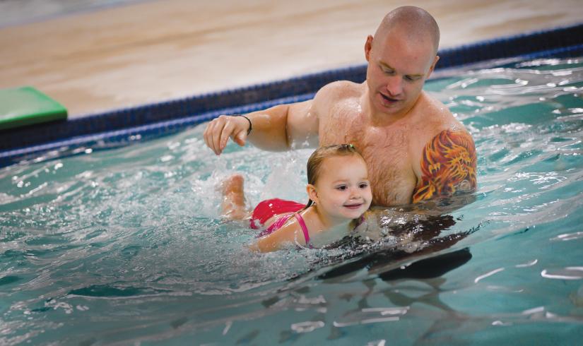 A father holds his young daughter up in the water as they leans on a kickboard and creates splashes behind her. 