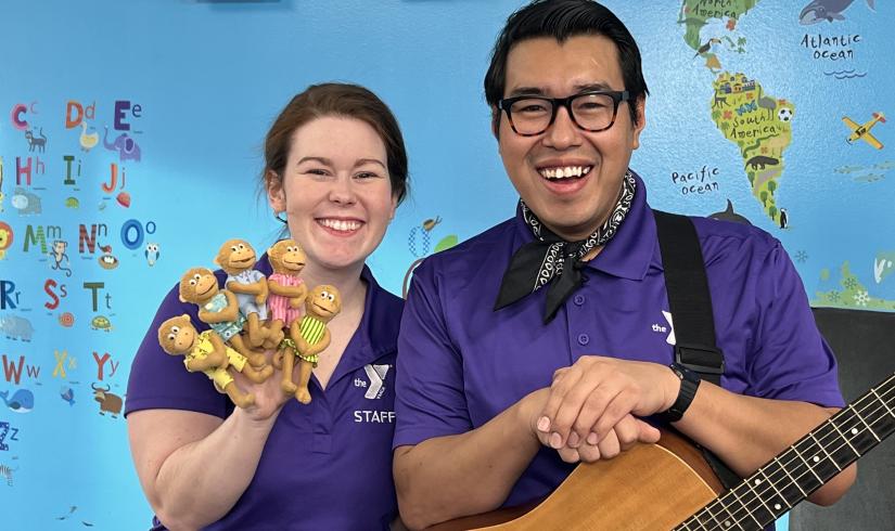YMCA360 instructors Lauren and Gabriel smile toward the camera in matching purple collared shirts. Lauren holds up 4 moneky finger puppets in pastel dresses. Gabriel wears a handkerchief and rests his hands on an acoustic guitar.