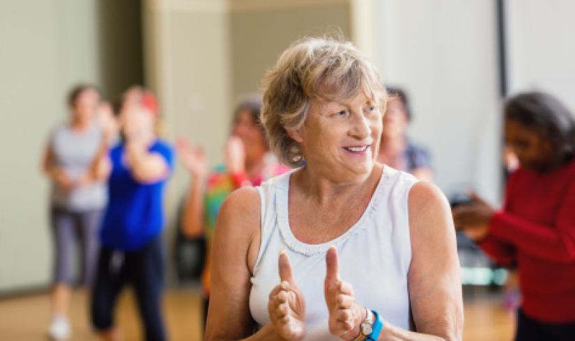 An active older woman in a white tank top and tawny shag haircut claps along in Enhance FItness class.
