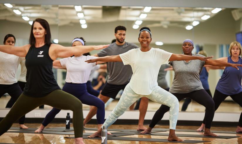 Women of different ages and ethnicities are in warrior pose in a YMCA studio with natural light.