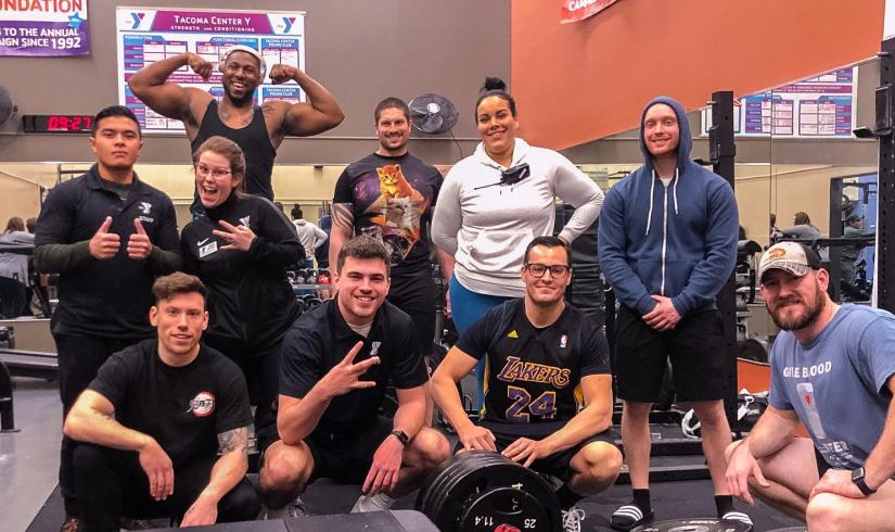 A group of adults huddle together and smile while inside the Tacoma Center YMCA weight room. 