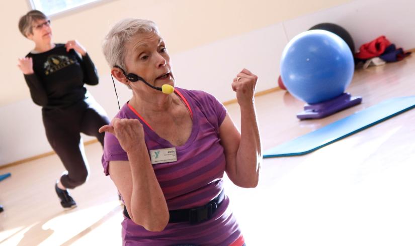 An active older instructor teaches core movement to a group of participants in a sun filled studio.