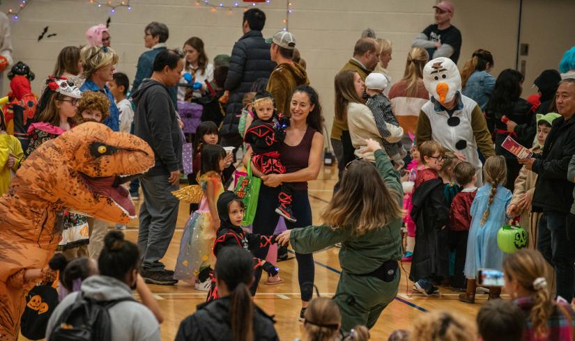 A crowd of adults and children are dressed up to celebrate the season. A raptor costume stands out in the forefront. 
