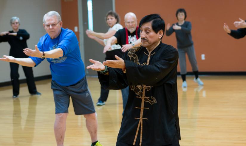 A group of older adults follows alongside and mirrors a Tai Chi Instructor in an all black outfit.