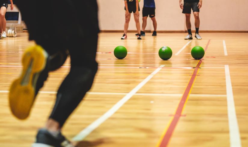A row of green dodgeballs line up in the center of a basketball court as a player runs up to grab one. 