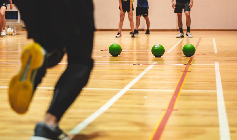 A row of green dodgeballs line up in the center of a basketball court as a player runs up to grab one.