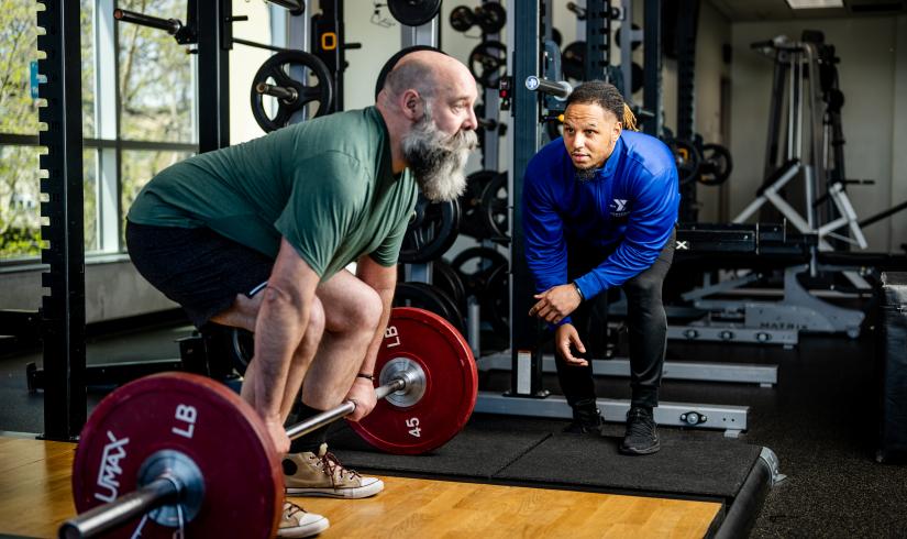 A man prepares to perform a deadlift with a Y staff member nearby in blue watching alongside for his form. 