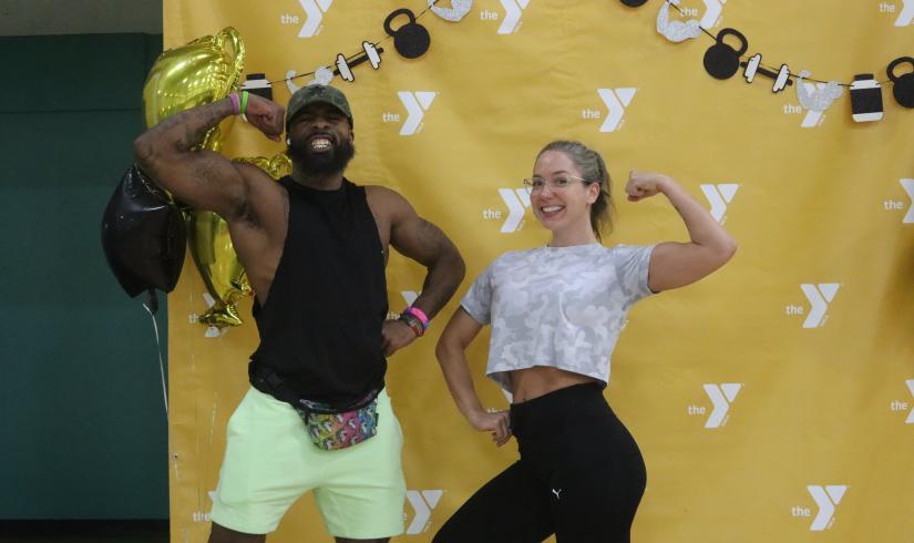 An African American man and a Caucasian woman pump their biceps in front of a yellow step and repeat with the Y logo.