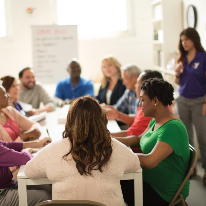 Diverse group of people sitting at a long table in the sunlight talking. Alongside them is a person near a white board with a YMCA polo shirt on.