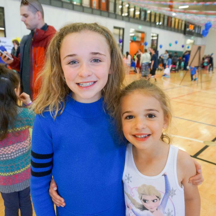 Two young girls stand side by side in a half hug within a glowing gymnasium. 