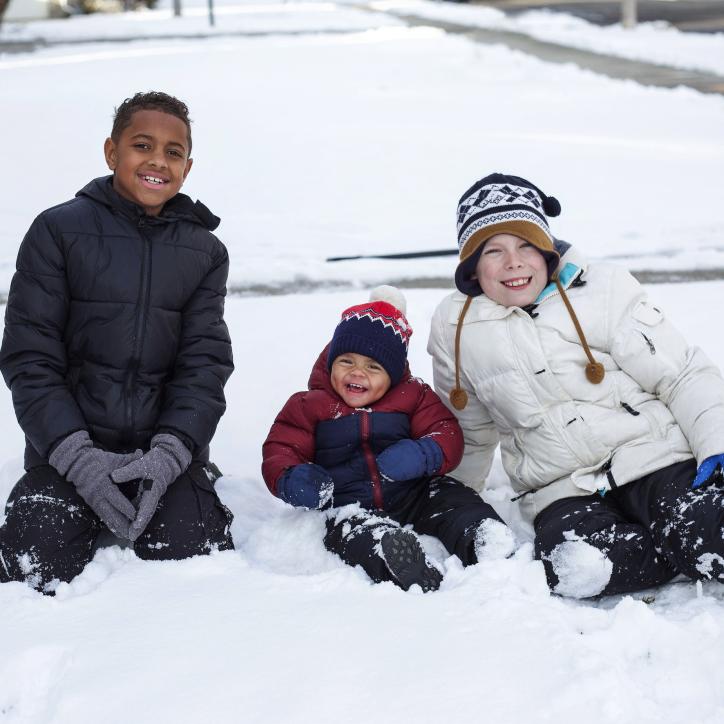 Three kids sit in the snow, two middle school age and one toddler.