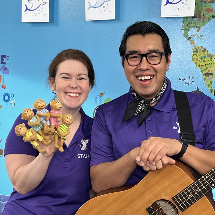 YMCA360 instructors Lauren and Gabriel smile toward the camera in matching purple collared shirts. Lauren holds up 4 moneky finger puppets in pastel dresses. Gabriel wears a handkerchief and rests his hands on an acoustic guitar.