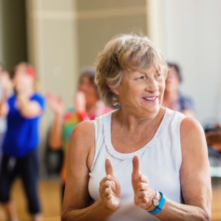 An active older woman in a white tank top and tawny shag haircut claps along in Enhance FItness class.