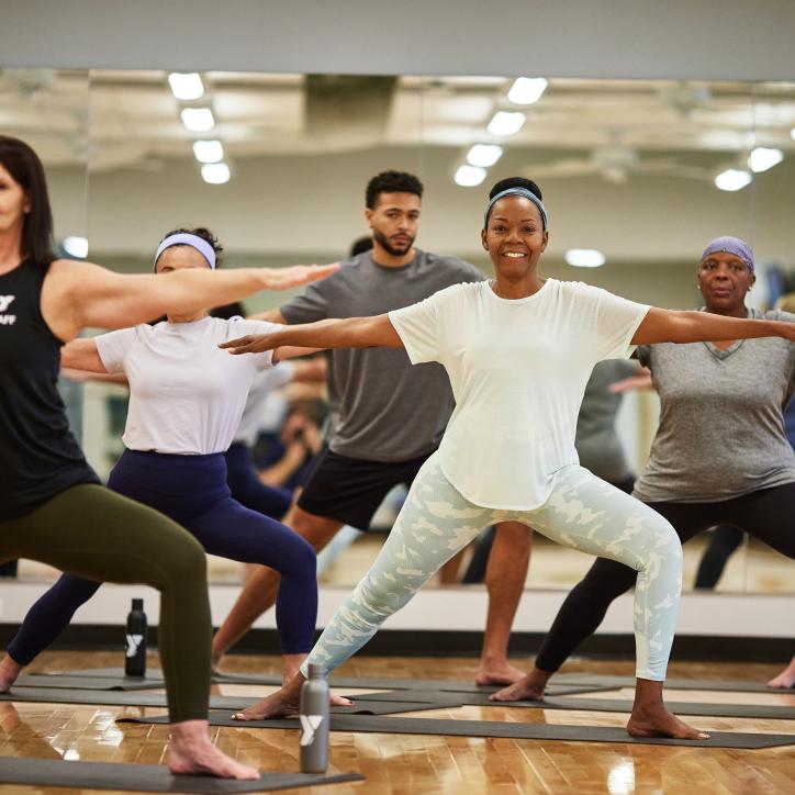 Women of different ages and ethnicities are in warrior pose in a YMCA studio with natural light.