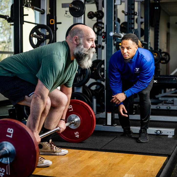 A man prepares to perform a deadlift with a Y staff member nearby in blue watching alongside for his form. 