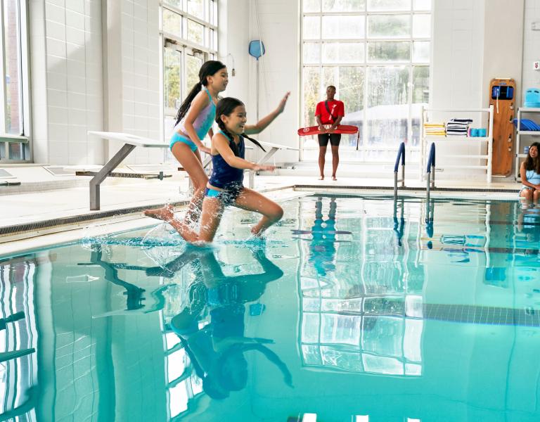 Kids Jumping In YMCA Pool With Lifeguard And Parent Watching