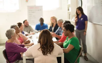 Diverse group of people sitting at a long table in the sunlight talking. Alongside them is a person near a white board with a YMCA polo shirt on.