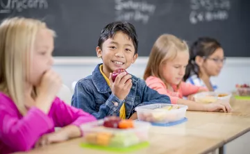 young boy smiling at the camera and holding an apple next to other young children sitting at a table