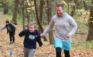 A father in son race up a trail, smiling as they move across fallen leaves on a cold day.