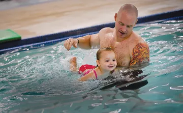 A father holds his young daughter up in the water as they leans on a kickboard and creates splashes behind her. 