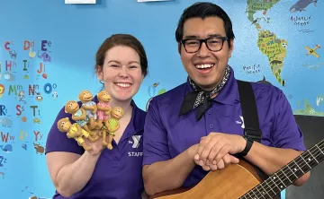 YMCA360 instructors Lauren and Gabriel smile toward the camera in matching purple collared shirts. Lauren holds up 4 moneky finger puppets in pastel dresses. Gabriel wears a handkerchief and rests his hands on an acoustic guitar.